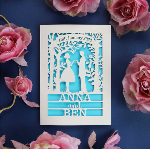 A personalised engagement card that features the silhouettes of a couple, and is personalised with a date in a banner at the top and the names of the couple over three lines of text at the bottom - A6 (small) / Peacock Blue