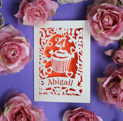 A personalised lasercut birthday card showing a cupcake with an age topper. Surrounded with heart, flowers and leaves, cut from cream card with an  orange insert. Personalise with name and age.