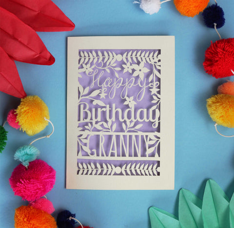 A personalised laser cut birthday card that says Happy Birthday with Granny underneath. Text is surrounded with flowers and leaves. This card is cream with a lilac paper backing. 