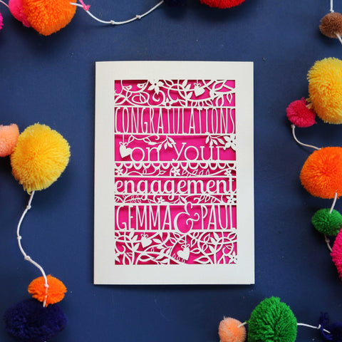 A personalised laser cut engagement card cut out of cream card and with a shocking pink paper behind. Card says "Congratulations on your engagement," with two names at the bottom. 
