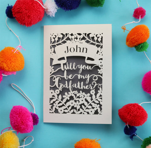 A personalised laser cut card with a banner containing the recipient's name, and a script font reading "Will you be my Godfather?". Card is laser cut from cream card and cut away to reveal a grey paper insert.