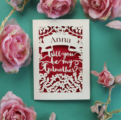 A personalised papercut card, laser cut from cream card and cut away to reveal a pink paper insert. Cut away design shows a banner with recipient's name and text underneath in a script font reads "Will you be my Godmother?"