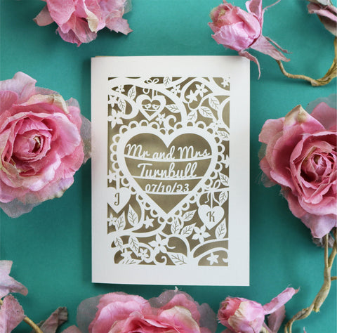 A laser cut wedding card personalised with the names and wedding date inside a laser cut heart border, surrounded by flowers and leaves. There are two smaller hearts with the initials of the bride and groom. 