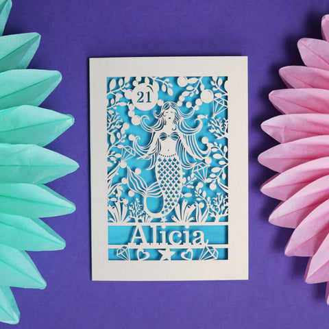 A personalised laser cut mermaid birthday card with a name and age. Cream card and a blue background.  - 