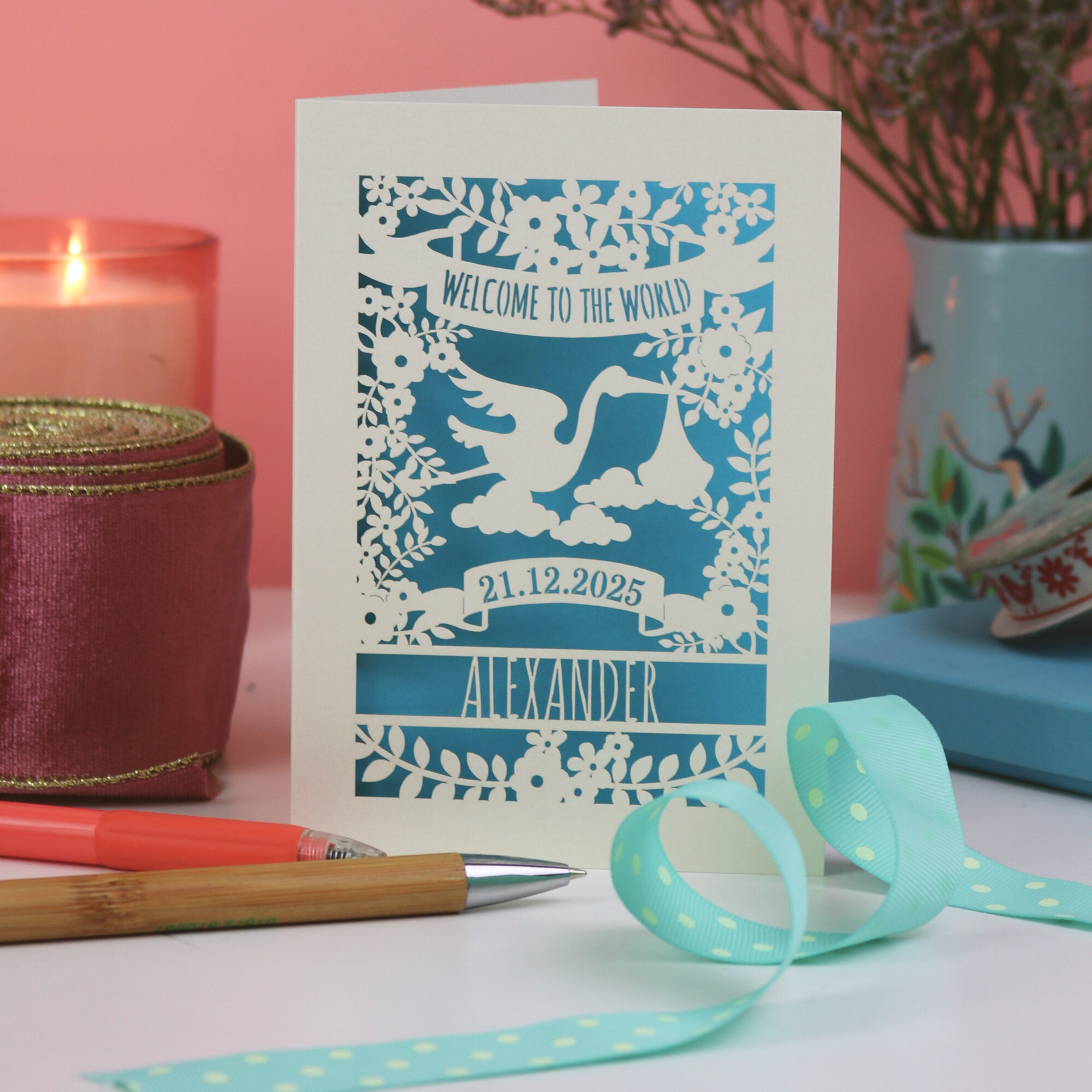 A laser cut New Baby card on pink background with plant, ribbon and candle