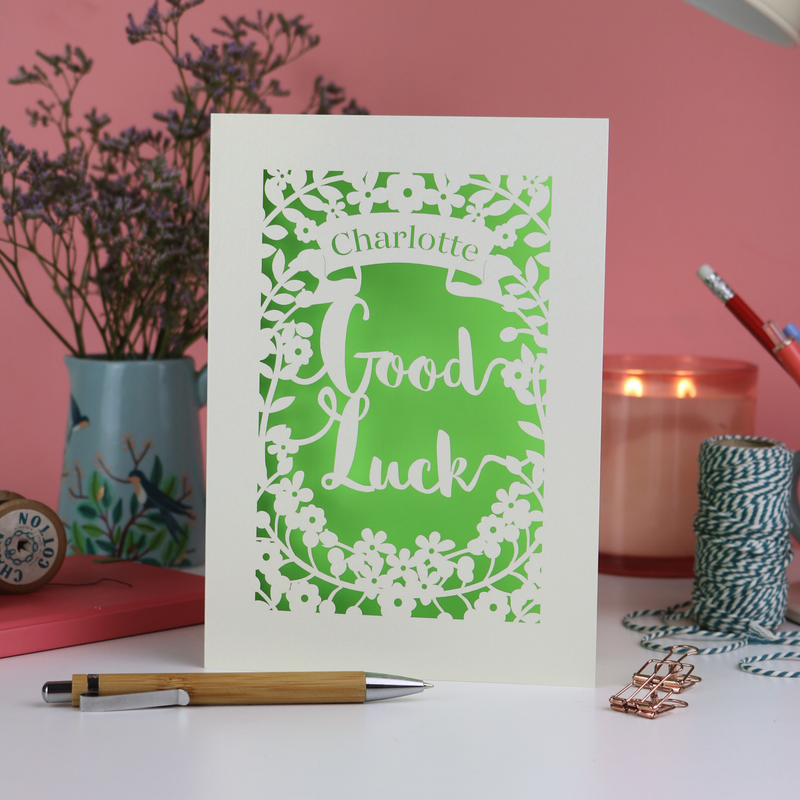 A photo of a laser cut good luck card on a pink background desk with plant, pens, twine and candle.