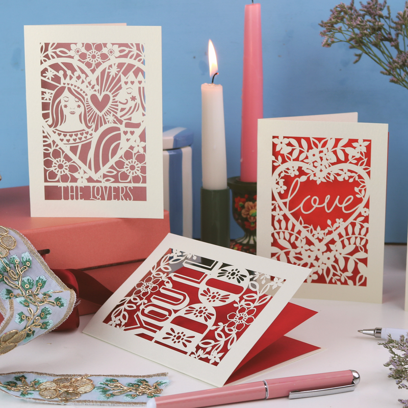 A collection of laser cut valentine's cards on a white desk with candles, pens and plants, on a blue background