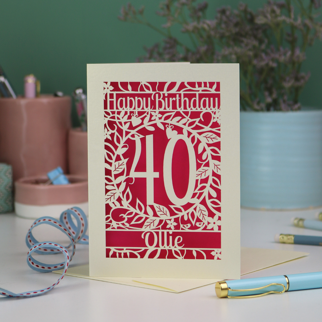A laser cut 40th birthday card personalised with a name and age, on a desk with stationery items 