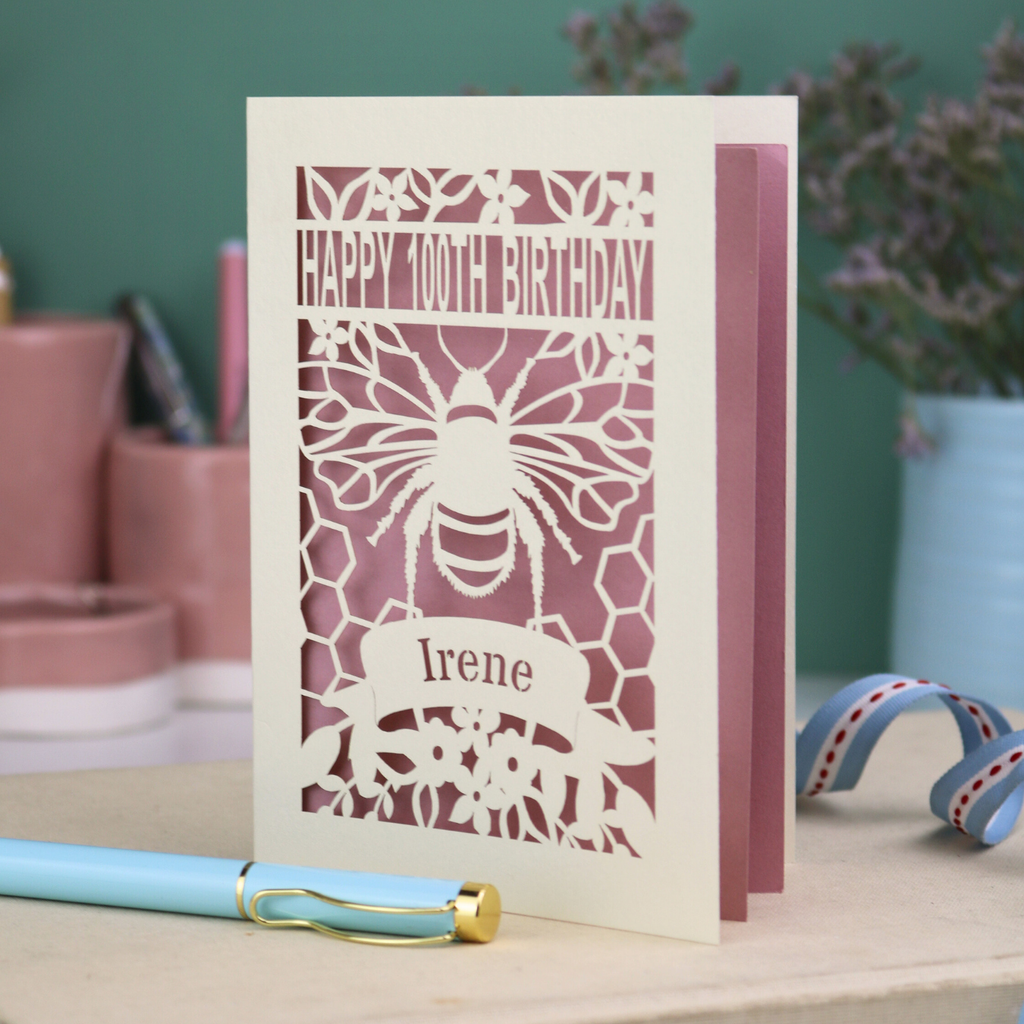 Laser-cut birthday card with bee design and 'Happy 100th Birthday Irene' text on a wooden surface.