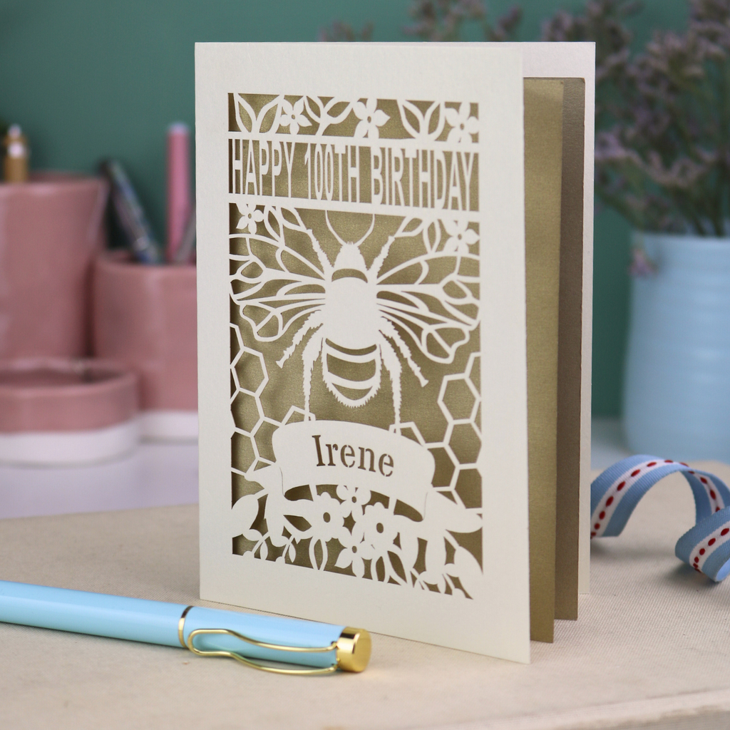 Laser-cut 100th birthday card with bee design and 'Happy 100th Birthday Irene' text on a table.