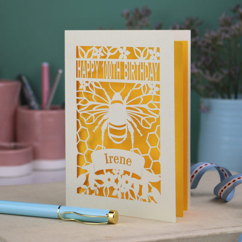 Birthday card with bee design and 'Happy 100th Birthday Irene' text on a wooden surface.