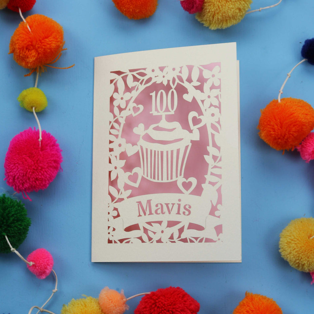 Decorative laser cut 100th card with '100 Mavis' design on a blue background with colourful pom-poms.