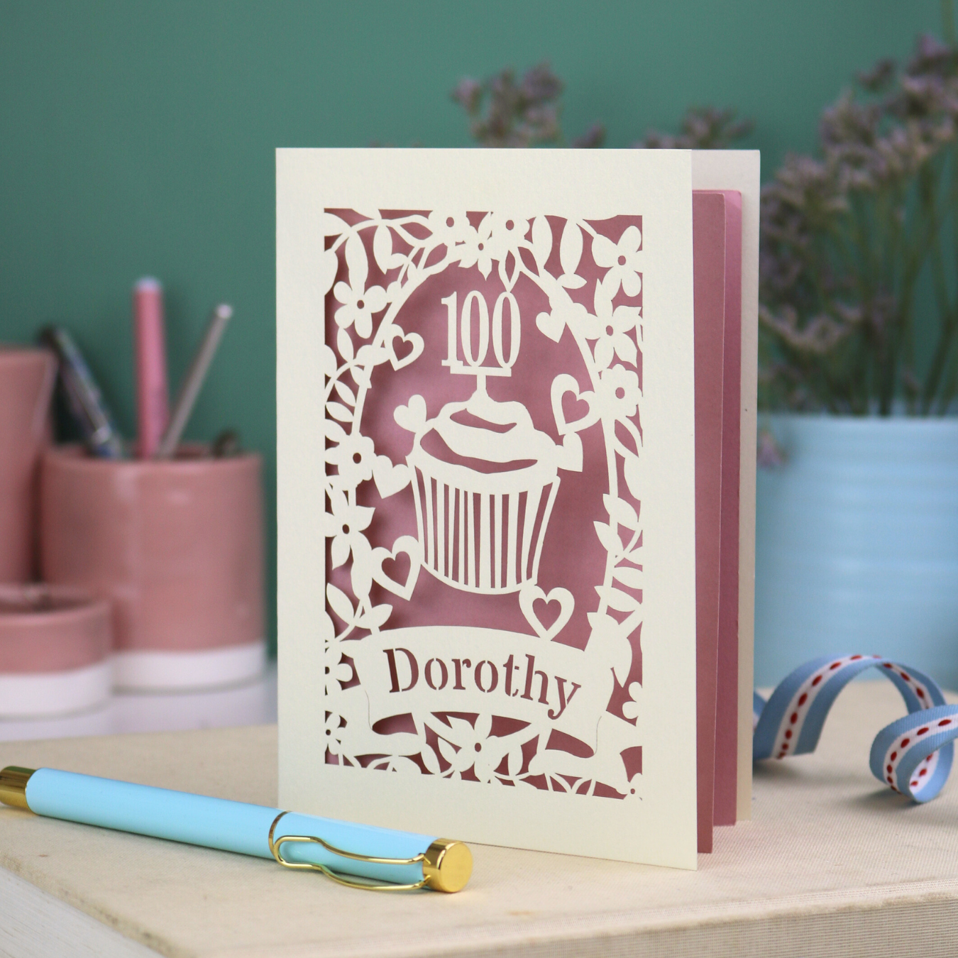 Decorative 100th birthday card with '100 Dorothy' cupcake and banner design on a table with stationery items.
