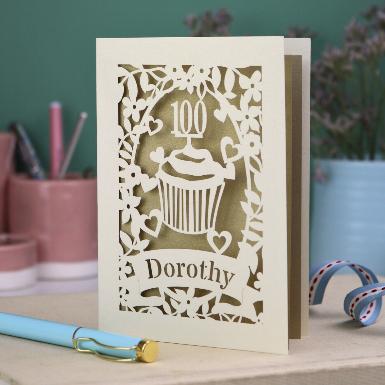 A laser cut 100th birthday card with cream floral cut out border, a cupcake with a 100 cake topper and a banner with the name "Dorothy" cut out in stencil font. 