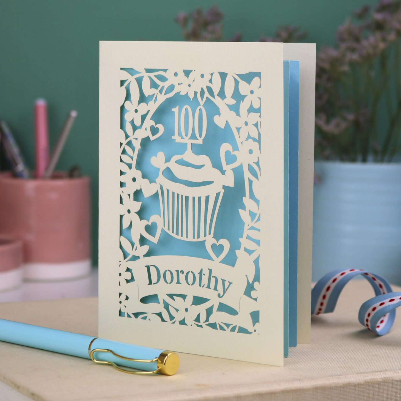 A laser cut 100th birthday card with cream floral cut out border, a cupcake with a 100 cake topper and a banner with the name "Dorothy" cut out in stencil font, and a light blue paper backing. 