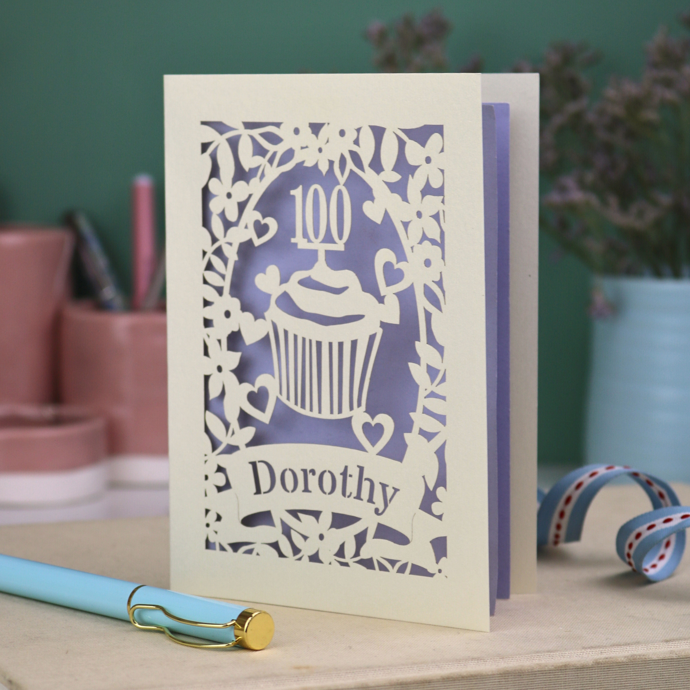 A laser cut 100th birthday card with cream floral cut out border, a cupcake with a 100 cake topper and a banner with the name "Dorothy" cut out in stencil font, and a lilac paper backing. 