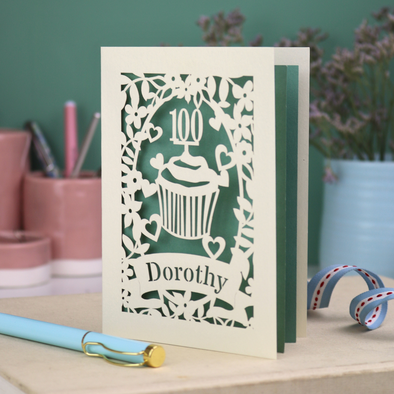 A laser cut 100th birthday card with floral cut out border, a cupcake with a 100 cake topper and a banner with the name "Dorothy" cut out in stencil font. 