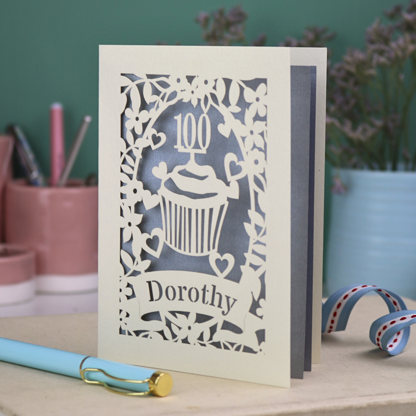 100 Birthday Card with decorative cut-out design featuring a cupcake and the number 100, with 'Dorothy' written below, on a desk with stationery items.