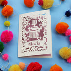laser cut 100th card with '100 Mavis' design on a blue background with colourful pom-poms.