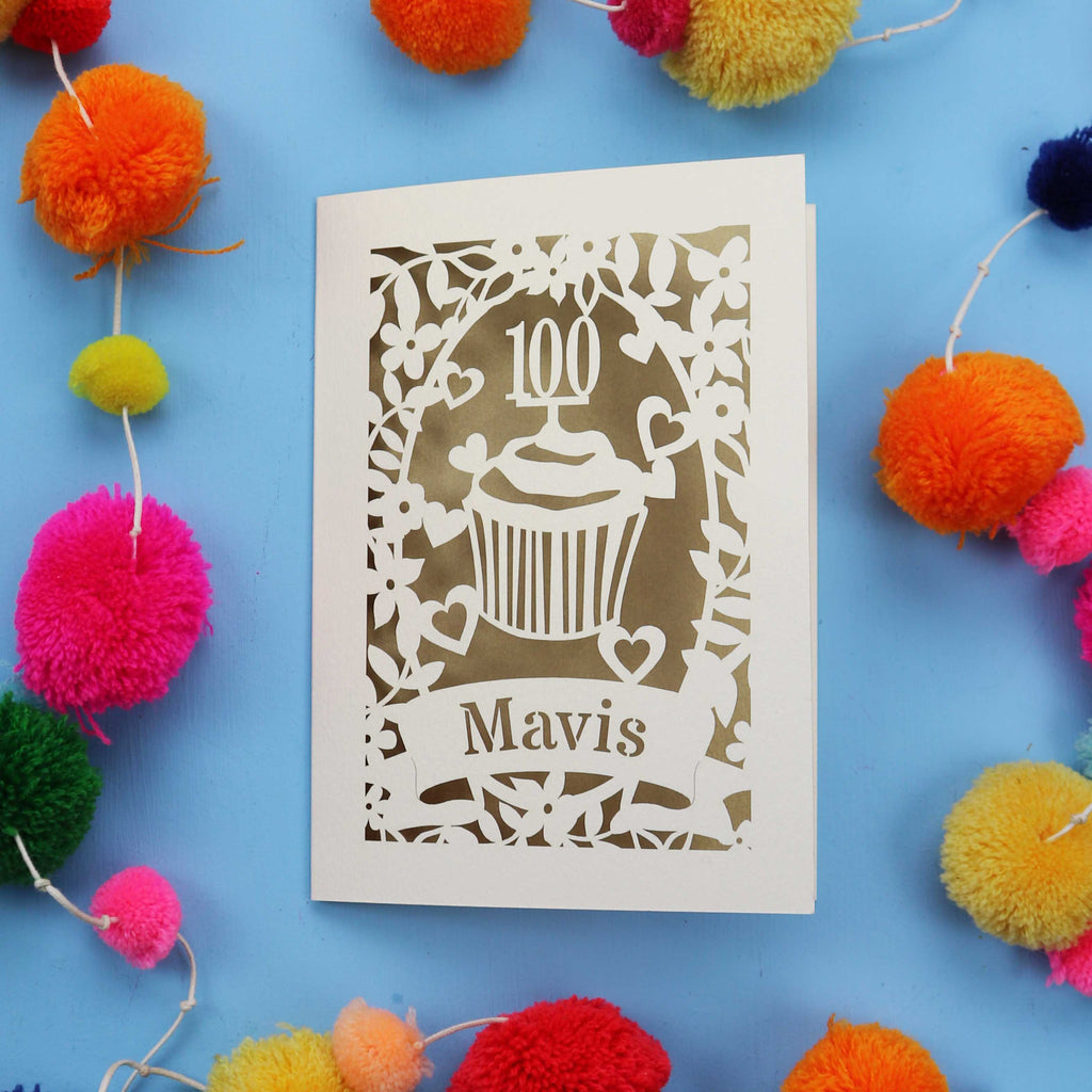 Decorative laser cut 100th card with '100 Mavis' design on a blue background with colourful pom-poms.