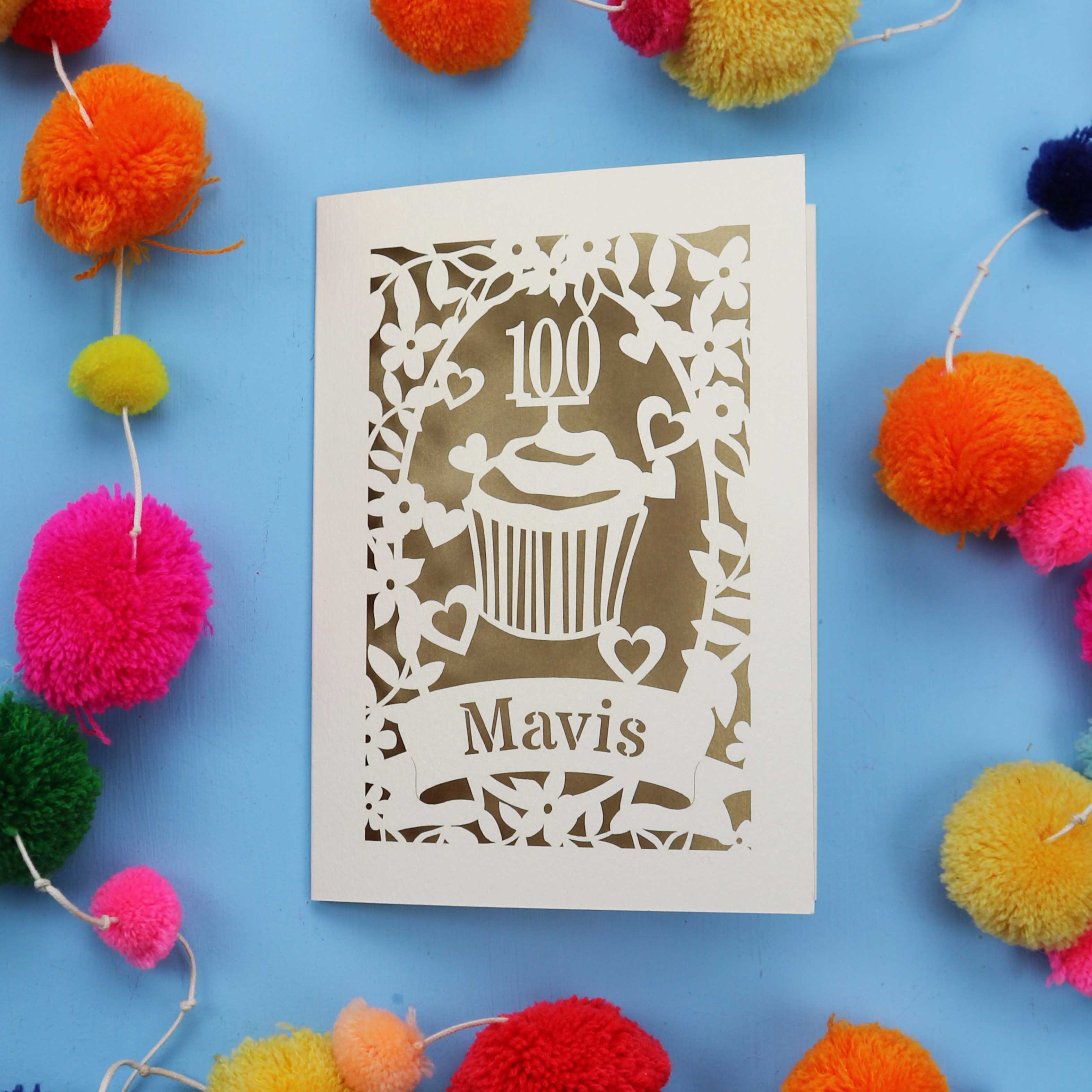 Decorative laser cut 100th card with '100 Mavis' design on a blue background with colourful pom-poms.