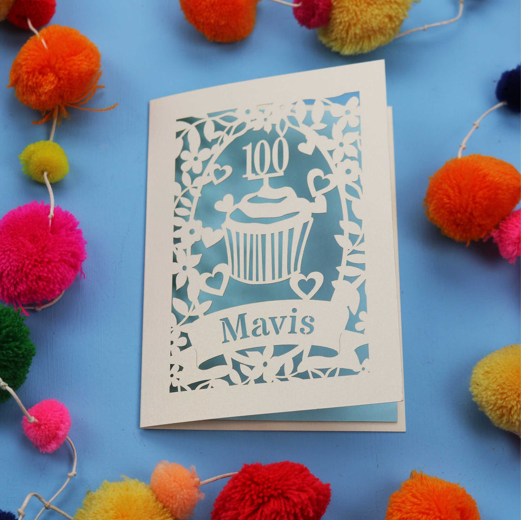 Decorative laser cut 100th card with '100 Mavis' design on a blue background with colourful pom-poms.
