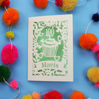 Decorative laser cut 100th card with '100 Mavis' design on a blue background with colourful pom-poms.
