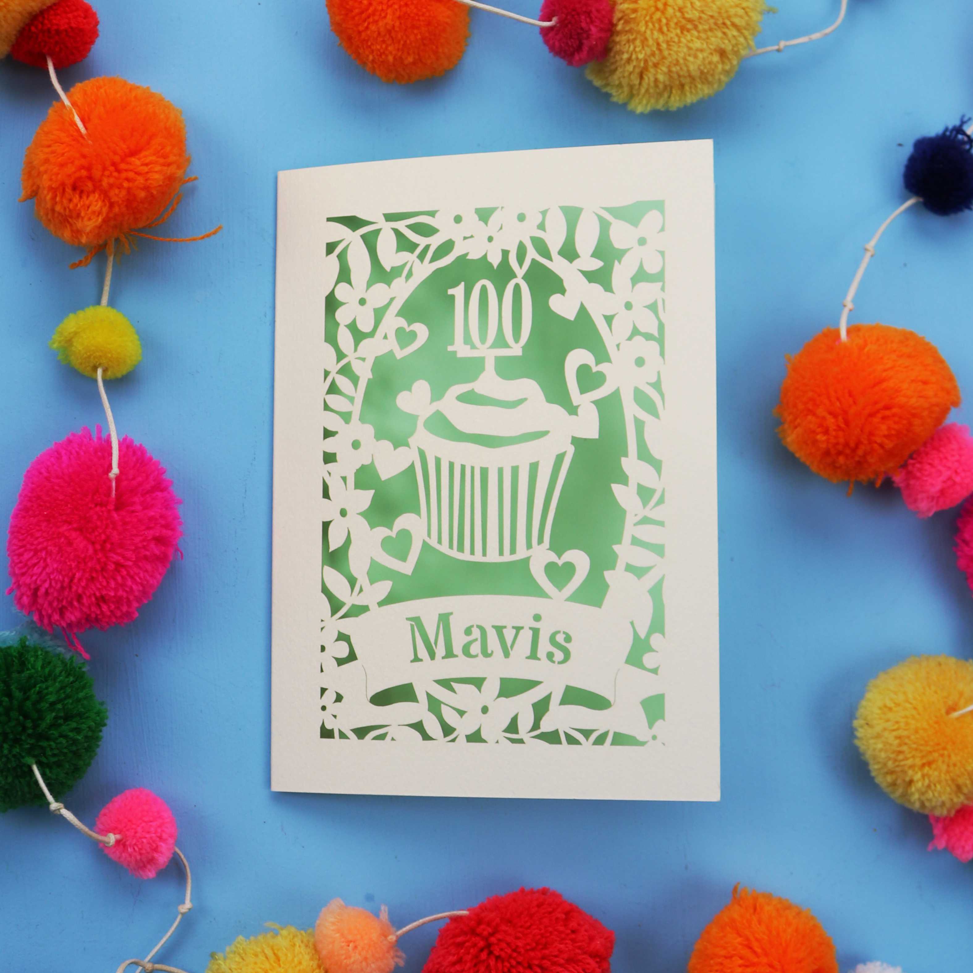 Decorative laser cut 100th card with '100 Mavis' design on a blue background with colourful pom-poms.