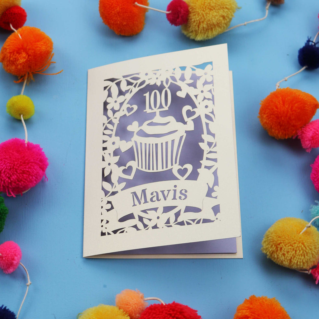 Decorative laser cut 100th card with '100 Mavis' design on a blue background with colourful pom-poms.