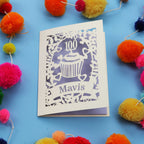 Decorative laser cut 100th card with '100 Mavis' design on a blue background with colourful pom-poms.
