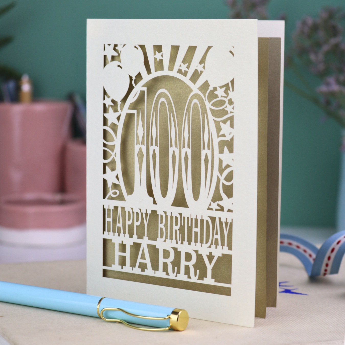 Personalized laser cut 100th birthday card with decorative cut out text, '100, Happy Birthday Harry' on a desk with stationery items.