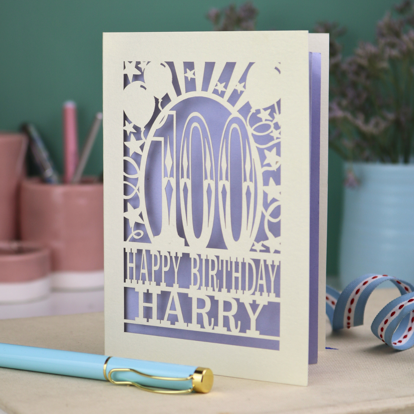 Personalized 100th birthday card with decorative cut out text, '100, Happy Birthday Harry' on a desk with stationery items.