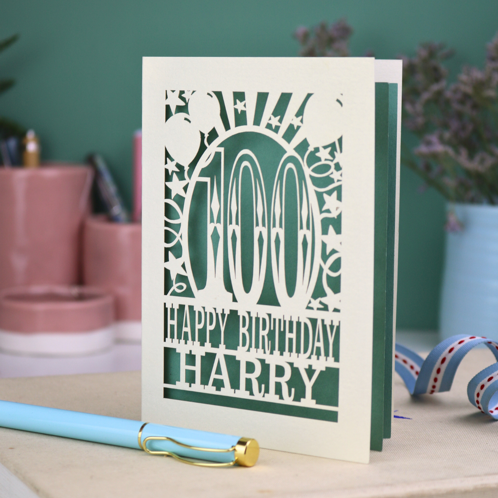 Personalized 100th birthday card with decorative cut out text, '100, Happy Birthday Harry' on a desk with stationery items.