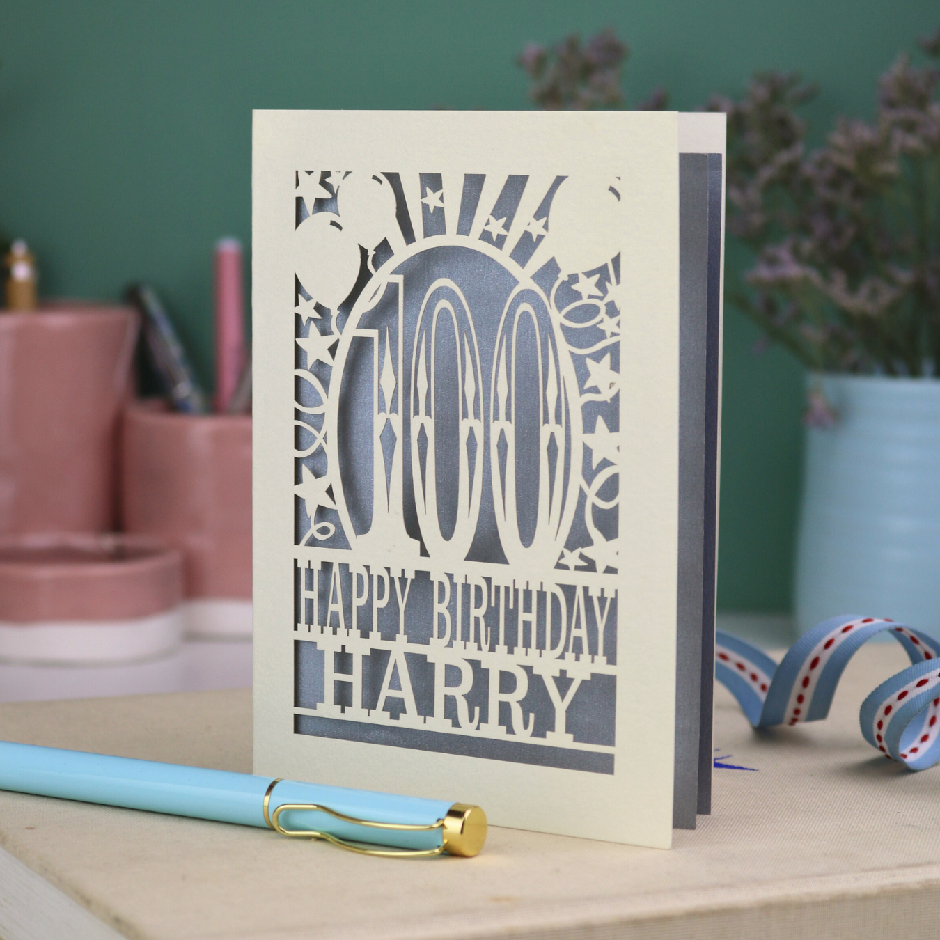 Personalized keepsake 100th birthday card with decorative cut out text, '100, Happy Birthday Harry' on a desk with stationery items.