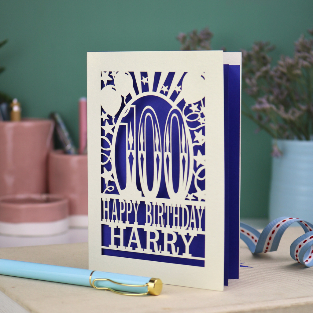 Personalised paper cut 100th birthday card with decorative cut out text, '100, Happy Birthday Harry' on a desk with stationery items.
