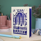 Personalised paper cut 100th birthday card with decorative cut out text, '100, Happy Birthday Harry' on a desk with stationery items.