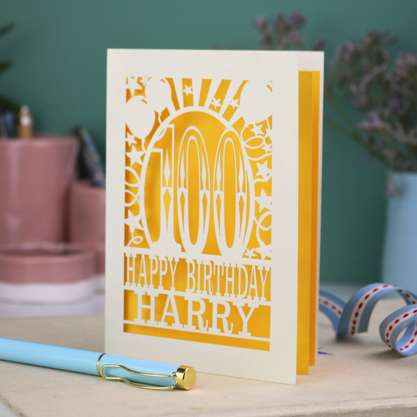 Personalized 100th birthday card with decorative cut out text, '100, Happy Birthday Harry' on a desk with stationery items.
