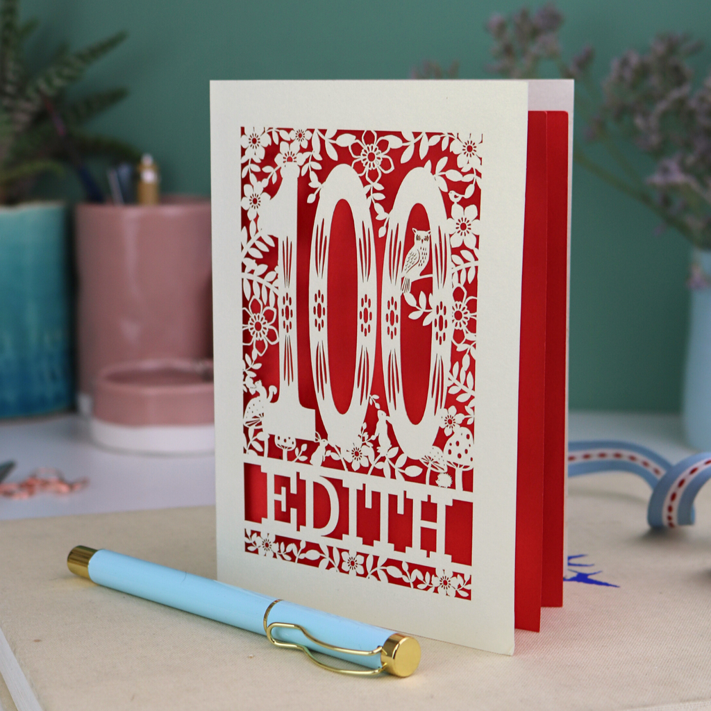 Laser cut 100th birthday Card with '100 Edith' design on a desk with a pen and plants in the background