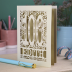 Laser cut 100th birthday Card with '100 Edith' design on a desk with a pen and plants in the background