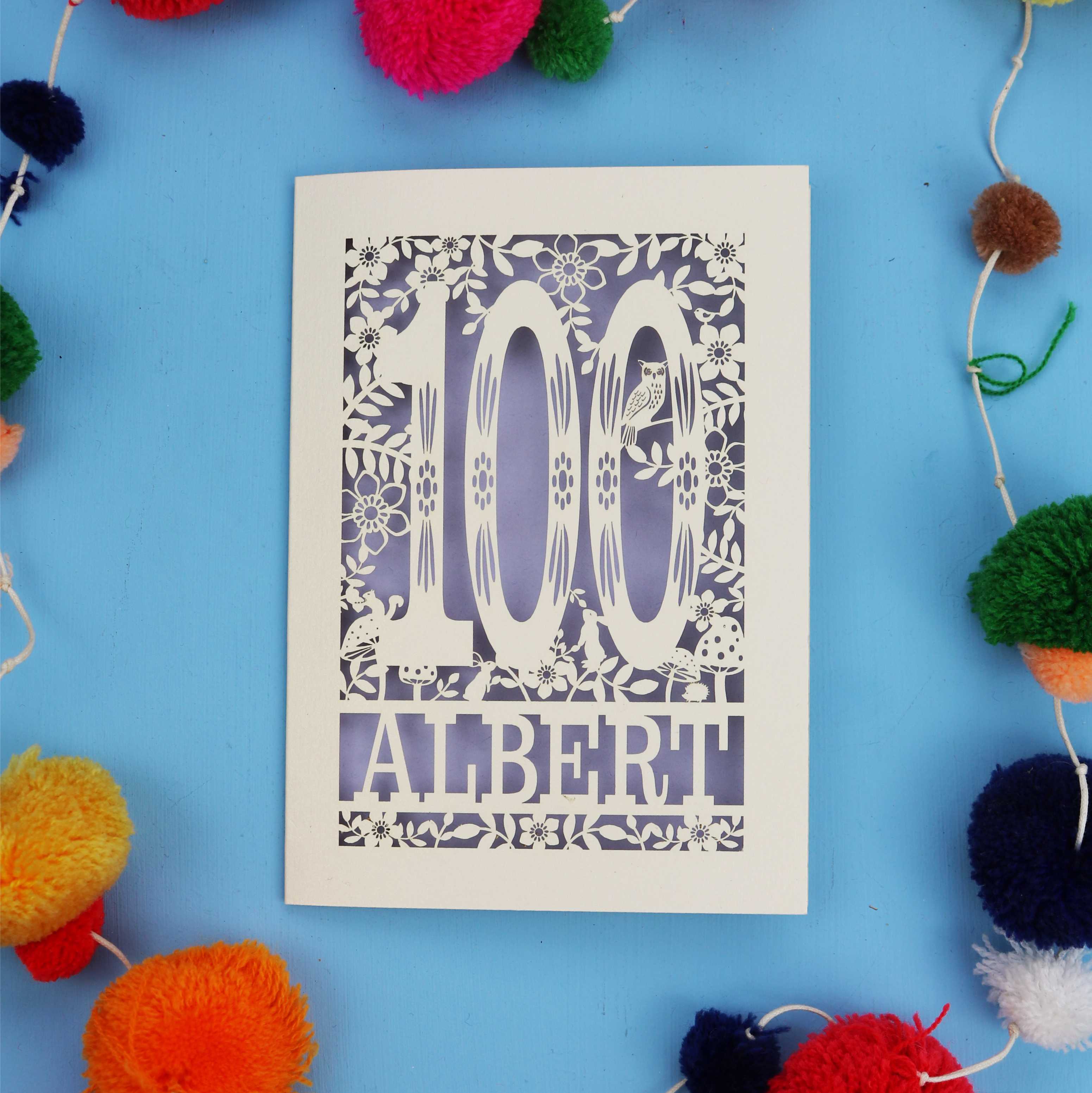 Decorative paper cut out 100th birthday card with '100 Albert' on a blue background with colourful pom-poms.