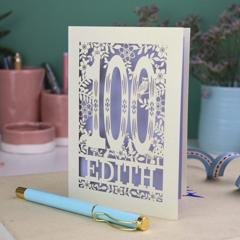 Laser cut 100th birthday Card with '100 Edith' design on a desk with a pen and plants in the background