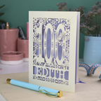 Laser cut 100th birthday Card with '100 Edith' design on a desk with a pen and plants in the background