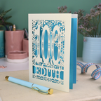 Laser cut 100th birthday Card with '100 Edith' design on a desk with a pen and plants in the background