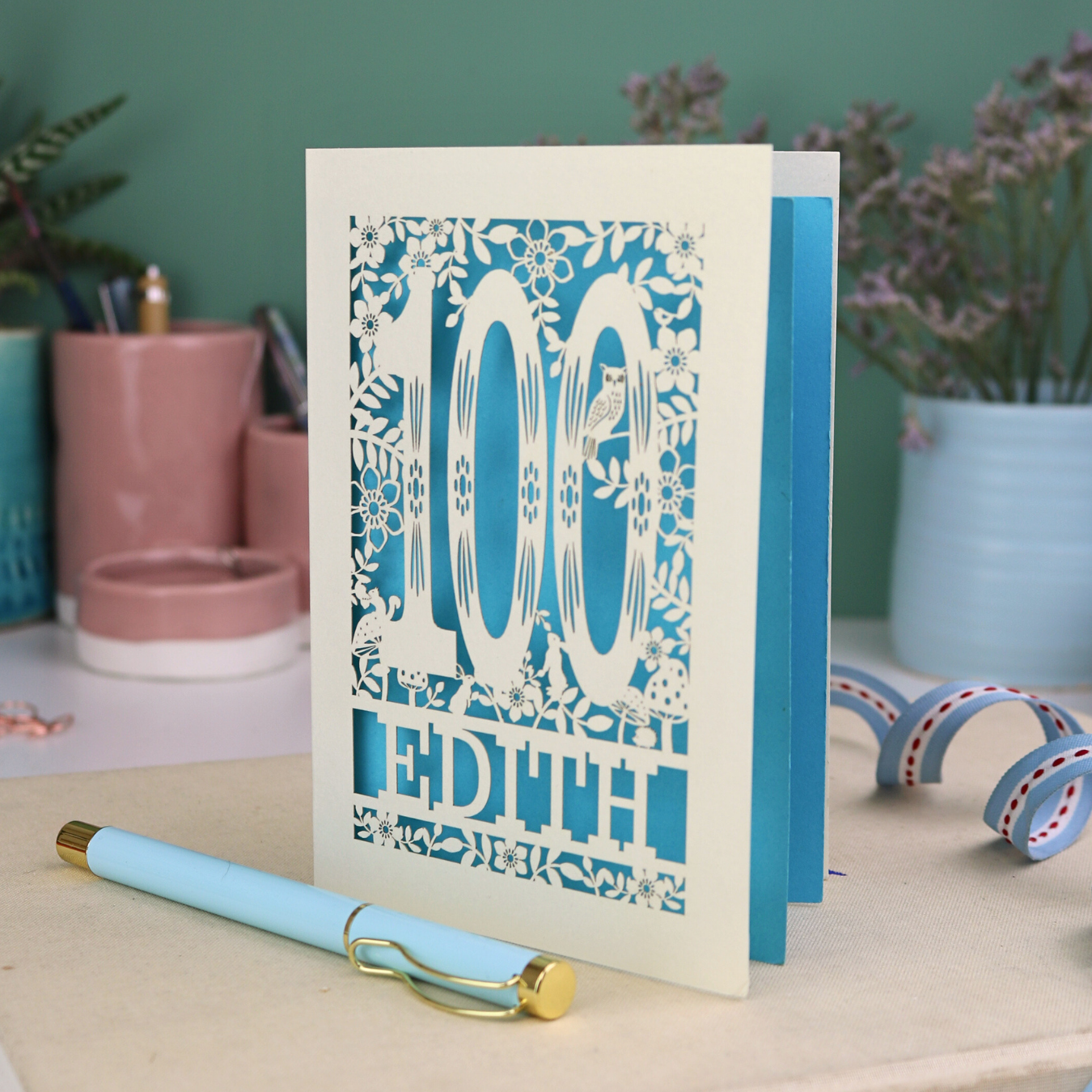 Laser cut 100th birthday Card with '100 Edith' design on a desk with a pen and plants in the background