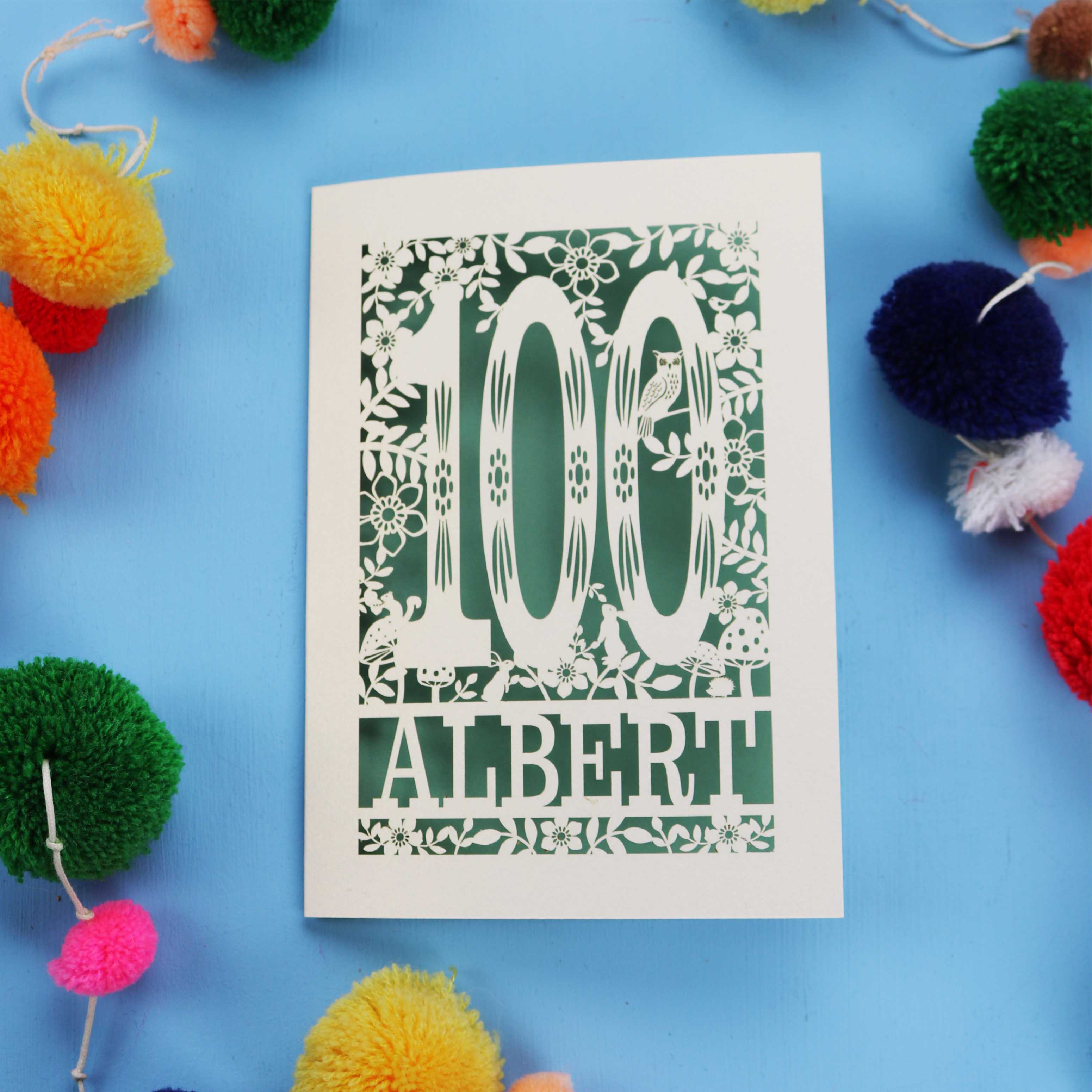 Decorative paper cut out 100th birthday card with '100 Albert' on a blue background with colourful pom-poms.
