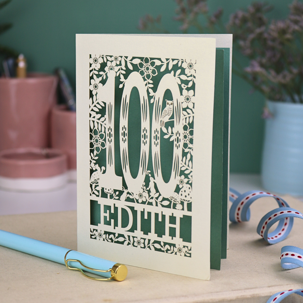 Laser cut 100th birthday Card with '100 Edith' woodland design on a desk with a pen and plants in the background
