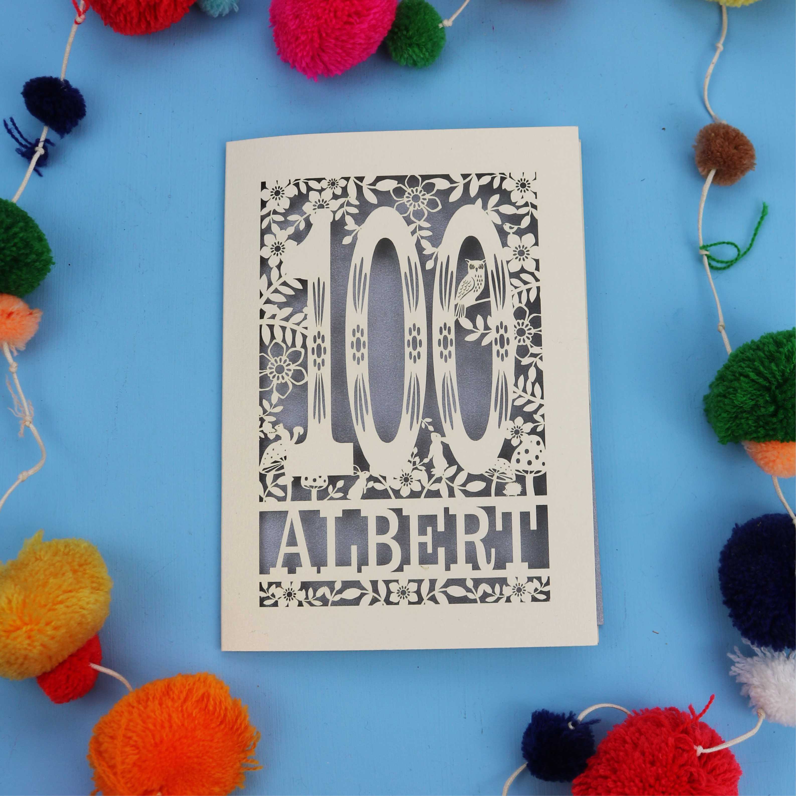 Decorative paper cut out 100th birthday card with '100 Albert' on a blue background with colourful pom-poms.