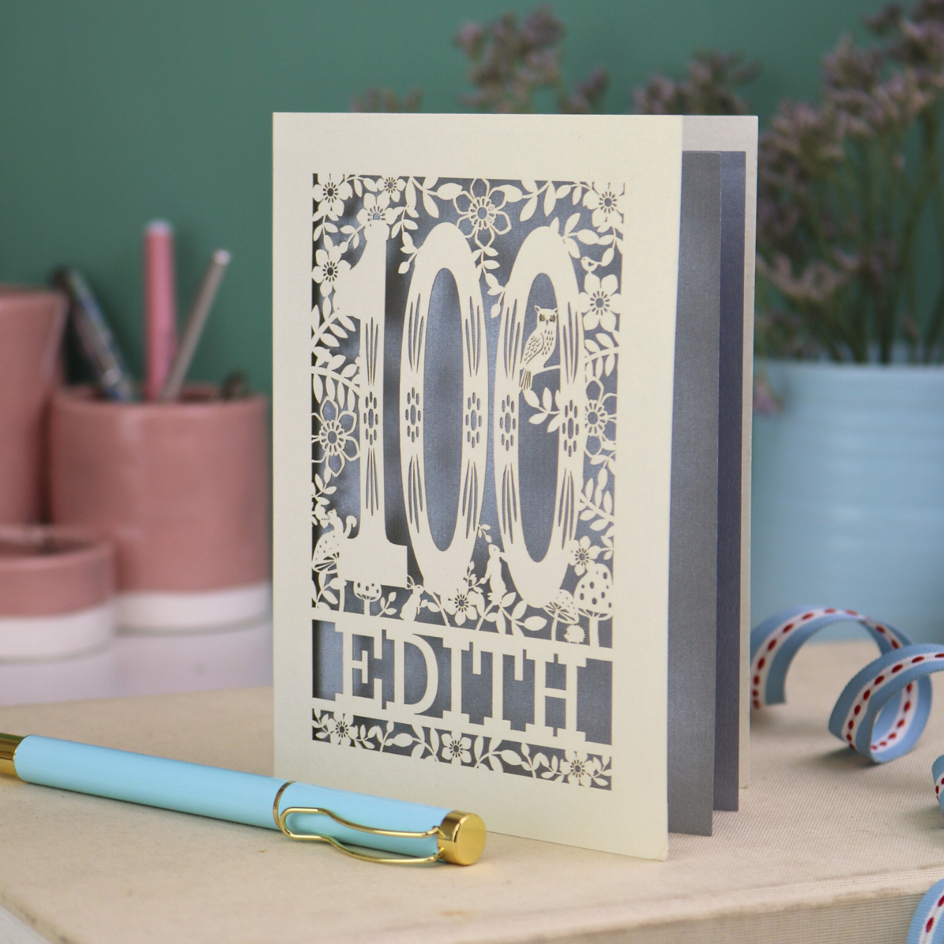 Laser cut 100th birthday Card with '100 Edith' design on a desk with a pen and plants in the background