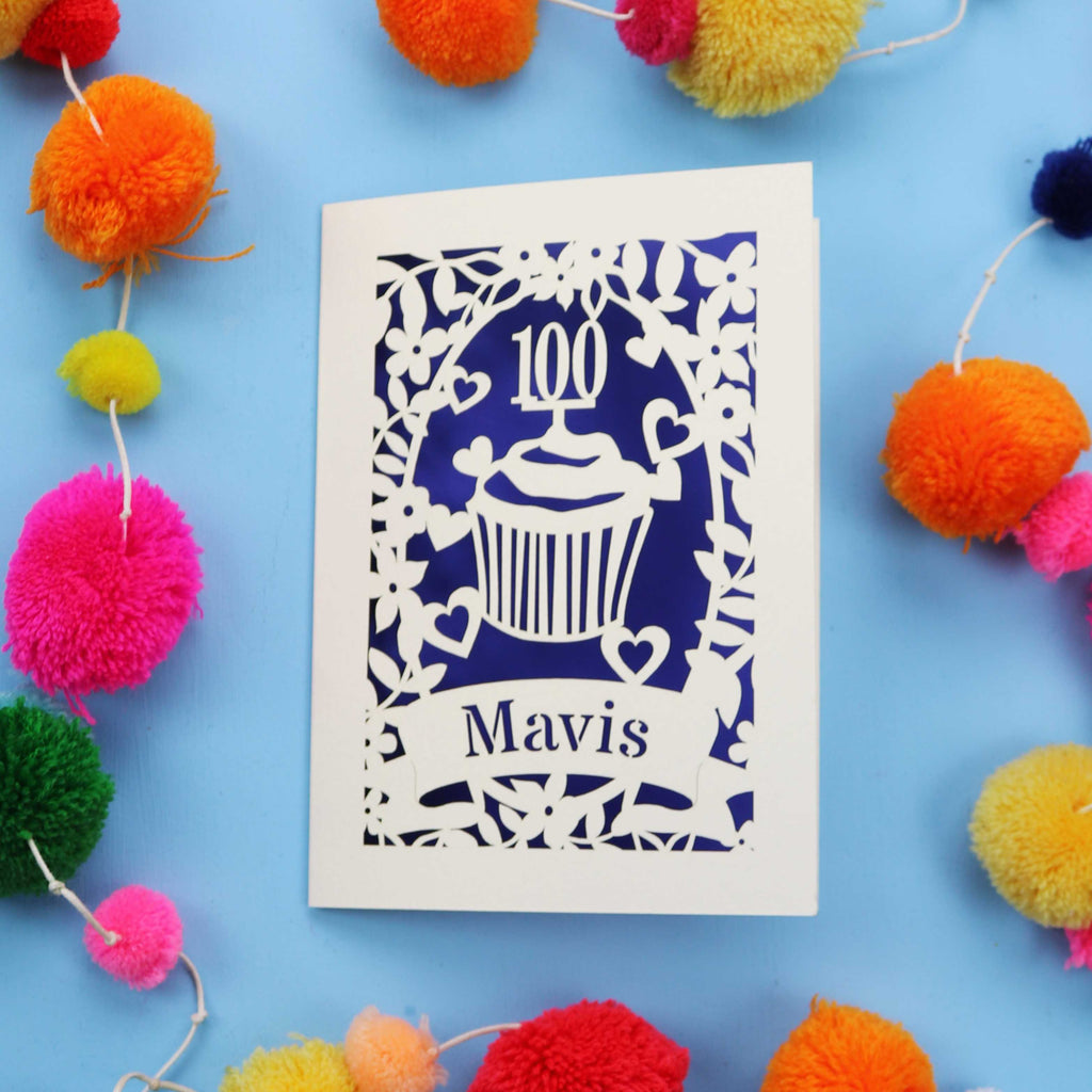 Personalised paper cut 100th card with '100 Mavis' design on a blue background with colourful pom-poms.