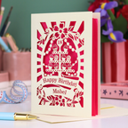 Laser-cut birthday card with pink and cream design on a desk with stationery items.